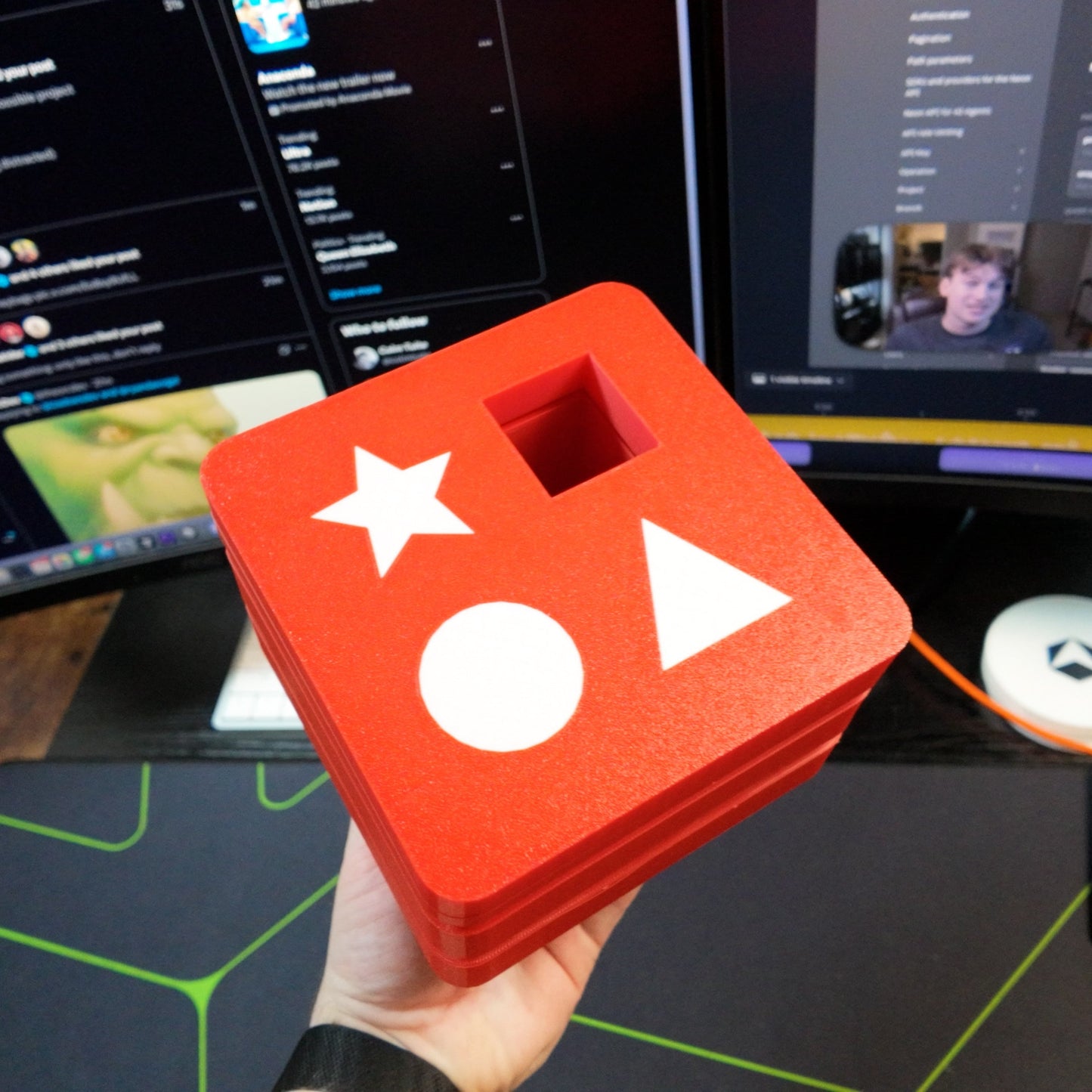 Red block with white stars and circles held in front of computer monitors displaying a video call.